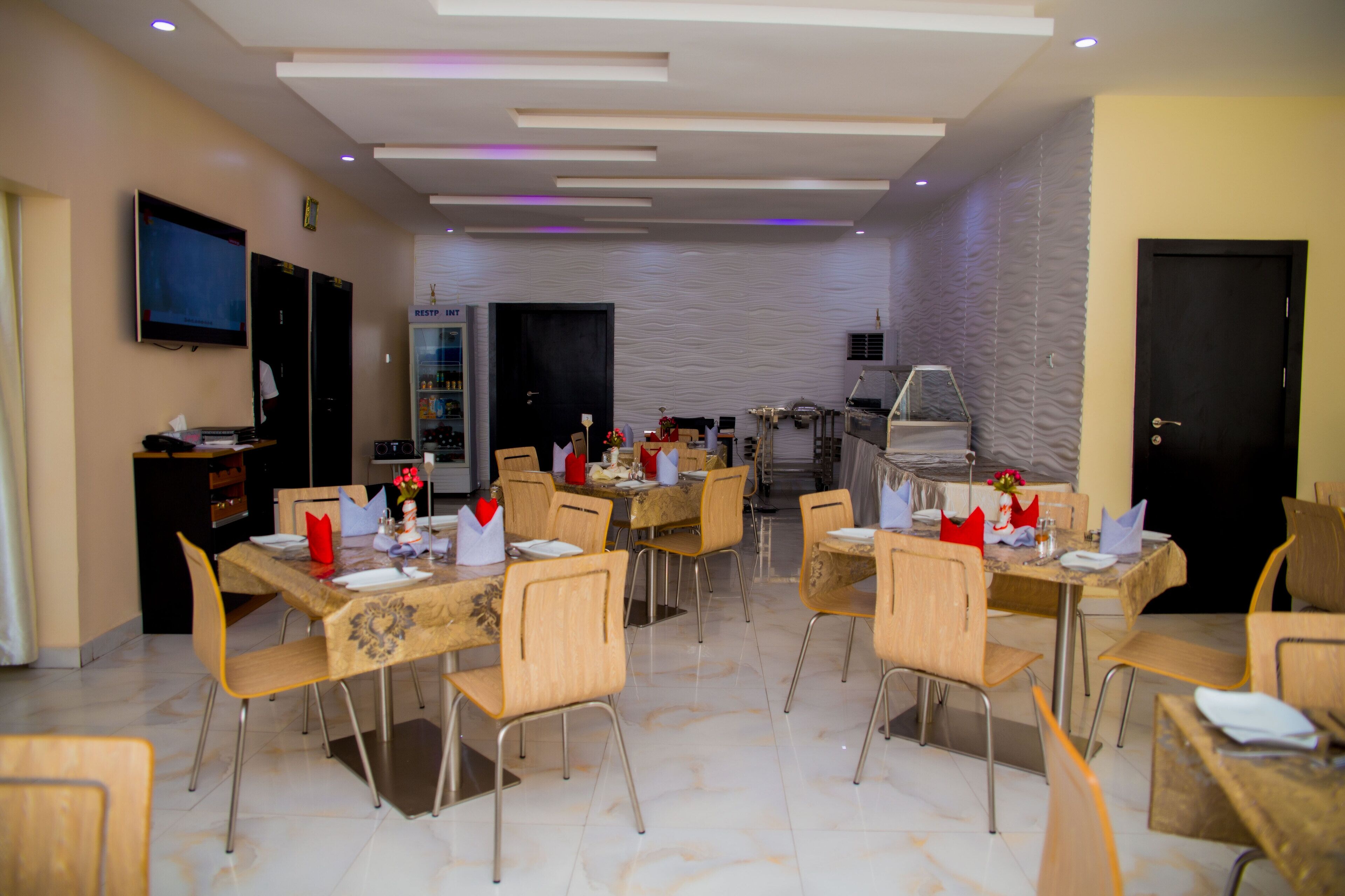 A modern dining area with multiple tables and chairs, a television mounted on the wall, and a kitchen area in the background.