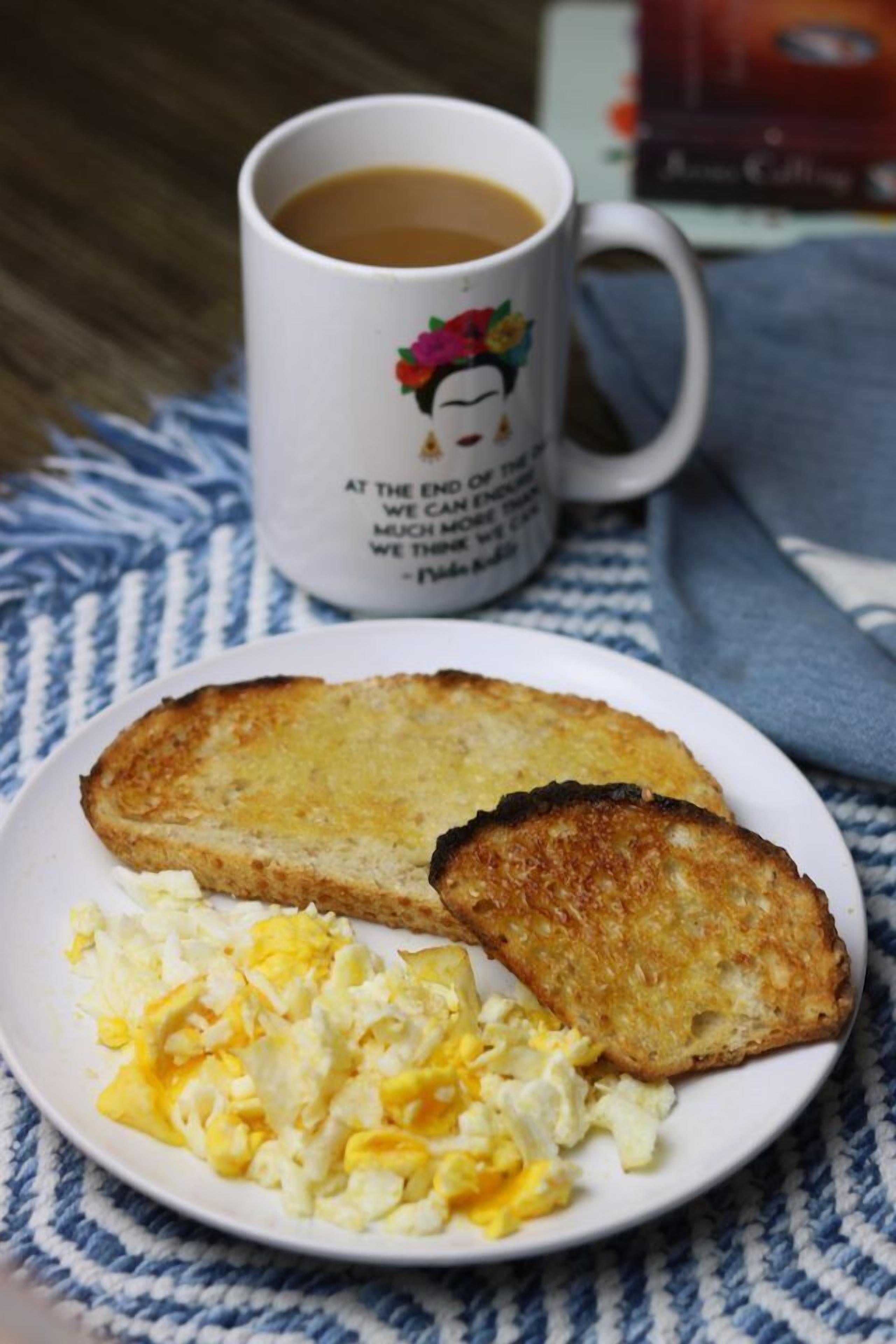 A plate with toast and scrambled eggs, a mug with a quote, and a book on a blue and white striped cloth.