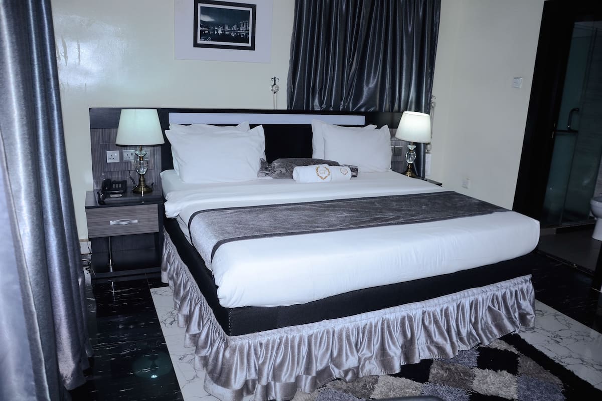 A neatly made bed with white and grey bedding, a black headboard, and a ruffled skirt. There are two bedside lamps, a nightstand with a phone, and a framed picture on the wall.