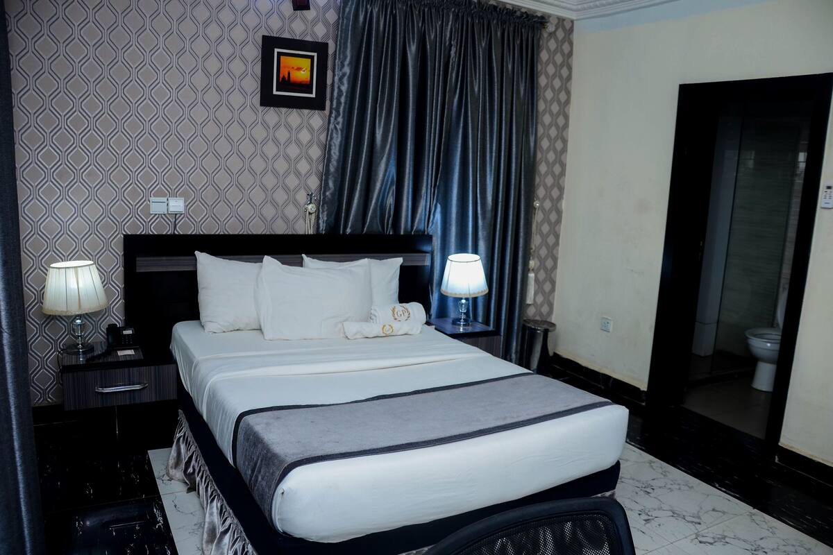 A hotel room with a neatly made bed, a bedside table with a lamp, a picture on the wall, and a bathroom door.
