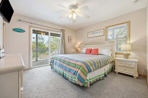 Une chambre à coucher comprenant un lit, une table de chevet, une commode, un ventilateur de plafond, une fenêtre avec des stores et une porte donnant sur un balcon.