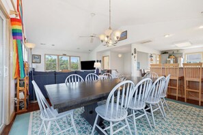 Un espace repas avec une grande table, des chaises blanches, un ventilateur de plafond et une vue sur l’extérieur.