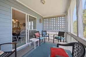 A covered porch with chairs and a table, a sliding glass door leading to a dining area, and a lattice railing. - 1712 - Happy Childhood, Corolla Coastal Home | Pool, Hot Tub & Game Room (Corolla)