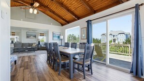 Un vaste espace de vie doté d’un plafond en bois, d’une table à manger avec des chaises, d’un canapé et d’une grande fenêtre offrant une vue sur les maisons.
