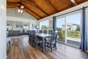 A spacious living area with a wooden ceiling, a dining table with chairs, a sofa, and a large window with a view of houses.