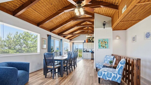 Une pièce spacieuse avec un plafond en bois, un coin repas avec des chaises, un canapé et une cuisine visible à travers une porte ouverte.