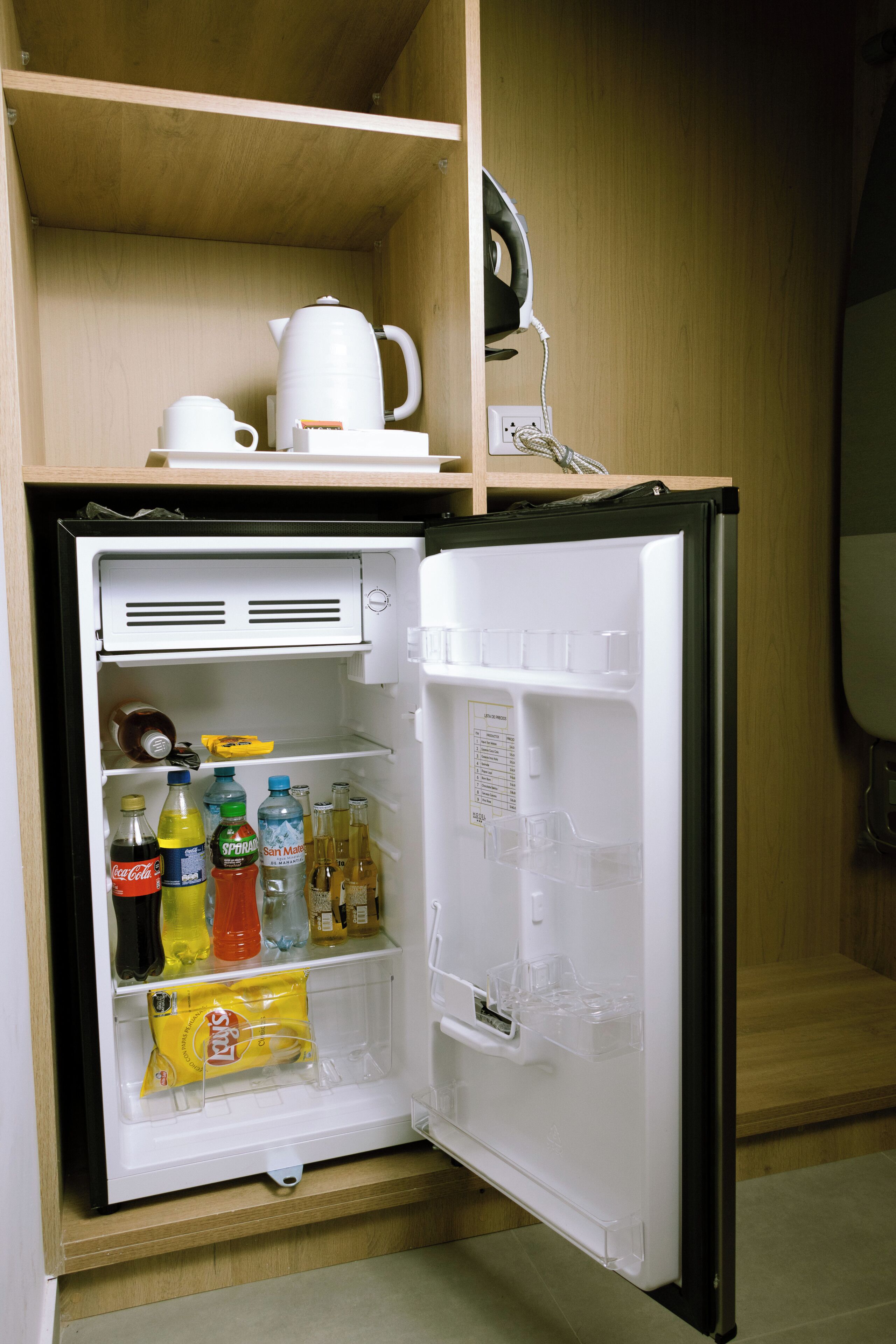 A compact refrigerator with a top shelf holding a white pitcher and a white mug. The door shelves contain various beverages and a yellow snack bag.