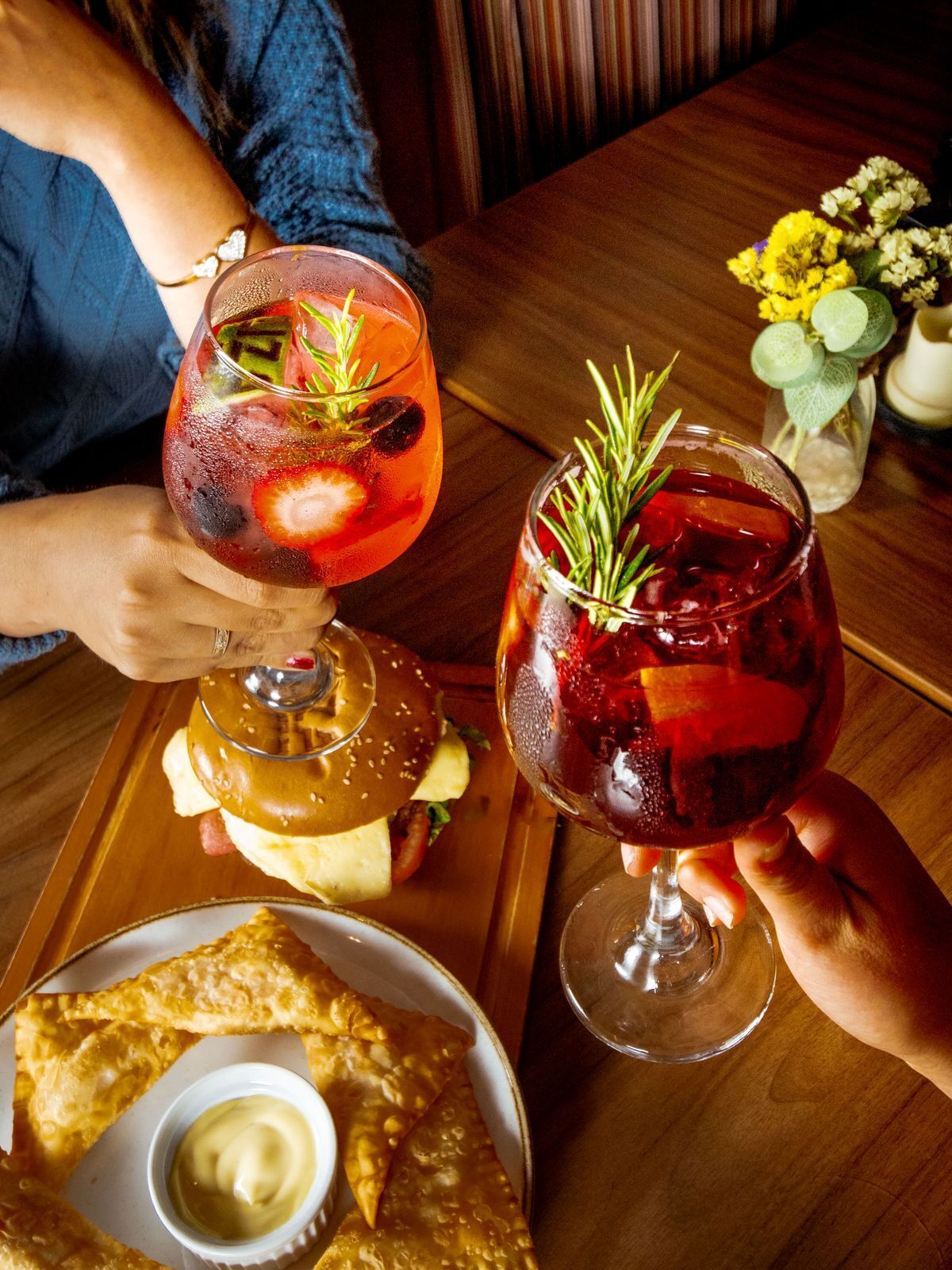 A dining table with a burger, a glass of red drink with ice and a sprig of rosemary, and a plate of fried items with dipping sauce.