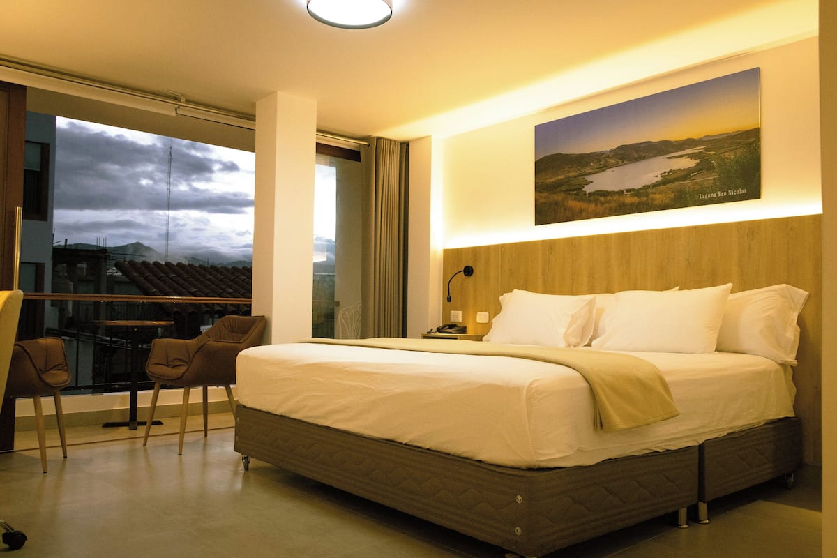 A modern hotel room with a large bed, a view of a cityscape through a large window, and a painting of a landscape on the wall.