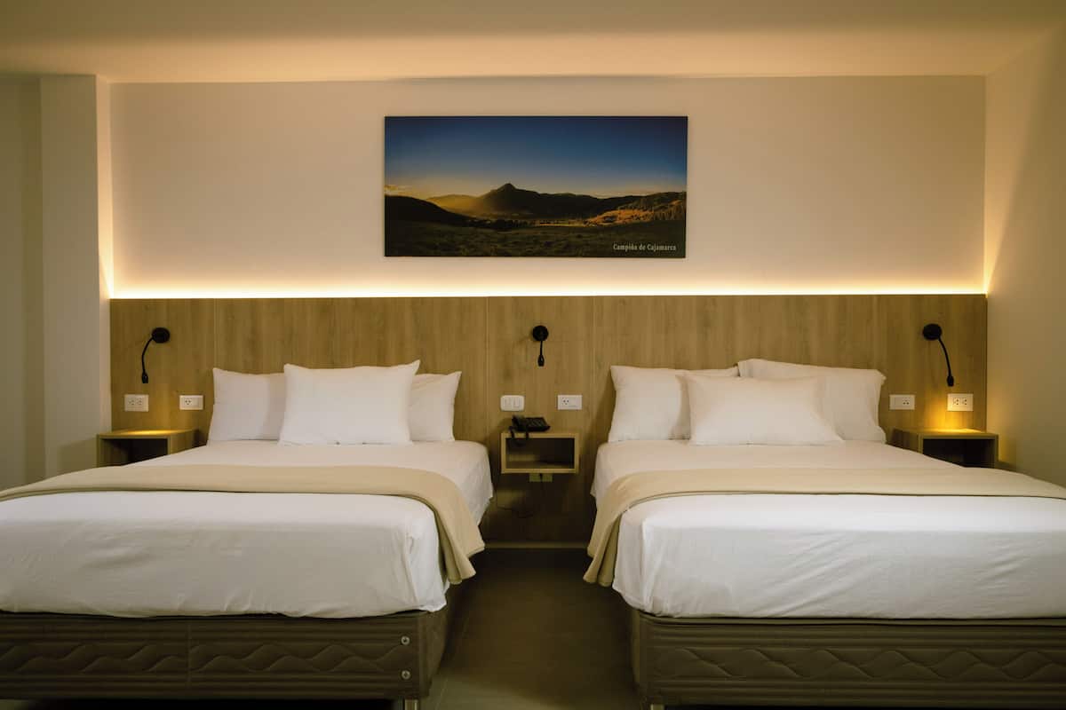 A hotel room with two beds, a wooden headboard, bedside lamps, and a framed landscape picture on the wall.