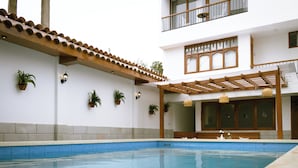 Una piscina con borde de azulejos, rodeada por un edificio blanco con una pérgola de madera y plantas en macetas.