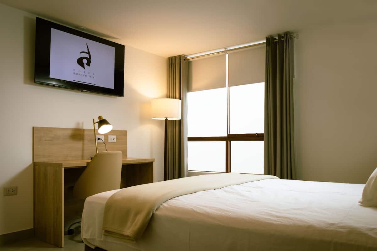 A hotel room with a bed, a desk, a chair, a TV mounted on the wall, and a window with curtains.