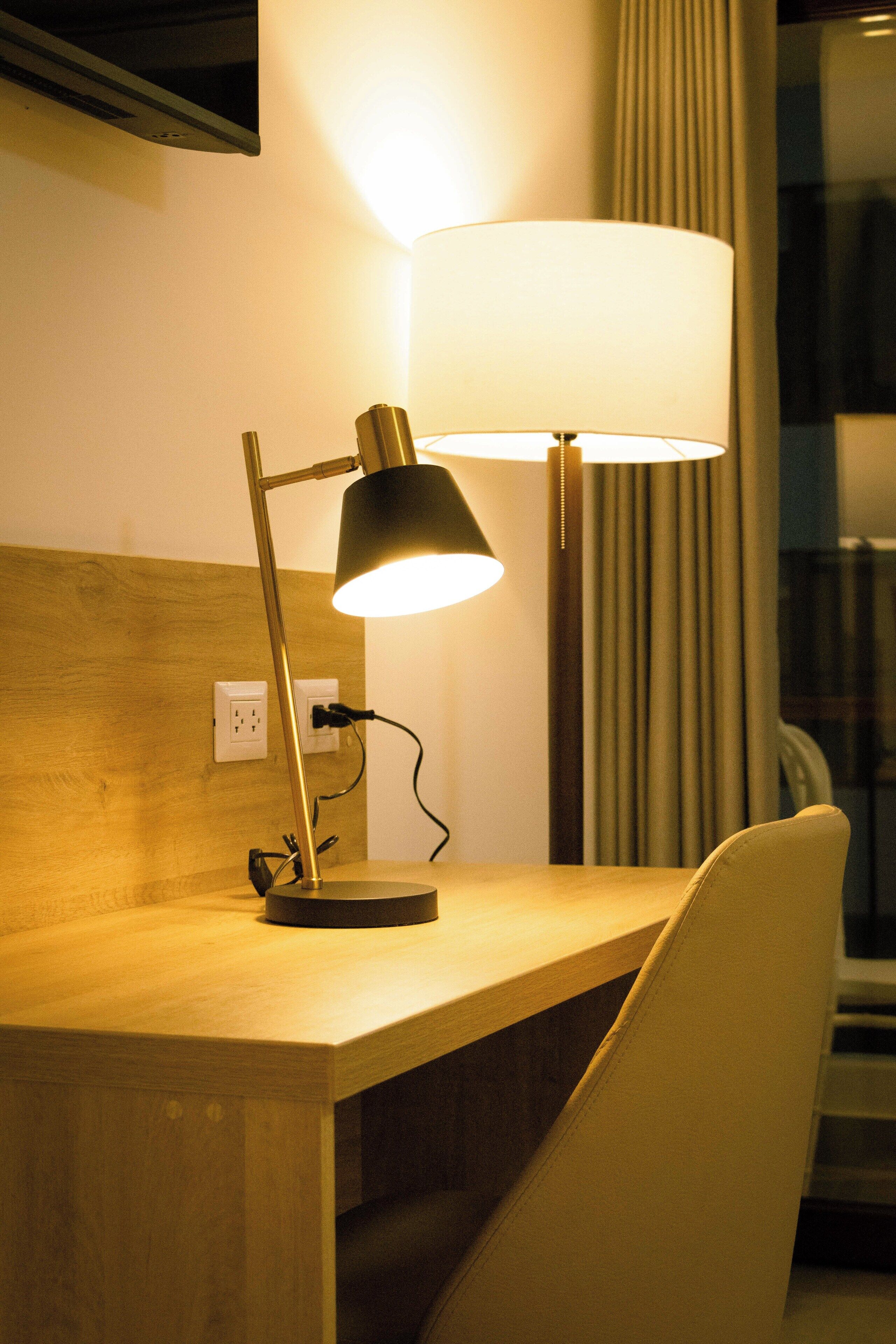 Ett skrivbord i trä med en lampa, en stol och ett fönster med gardiner.