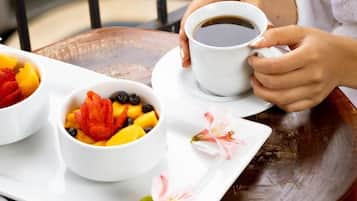 A person holding a cup of coffee, seated at a wooden table with bowls of fruit and a flower.