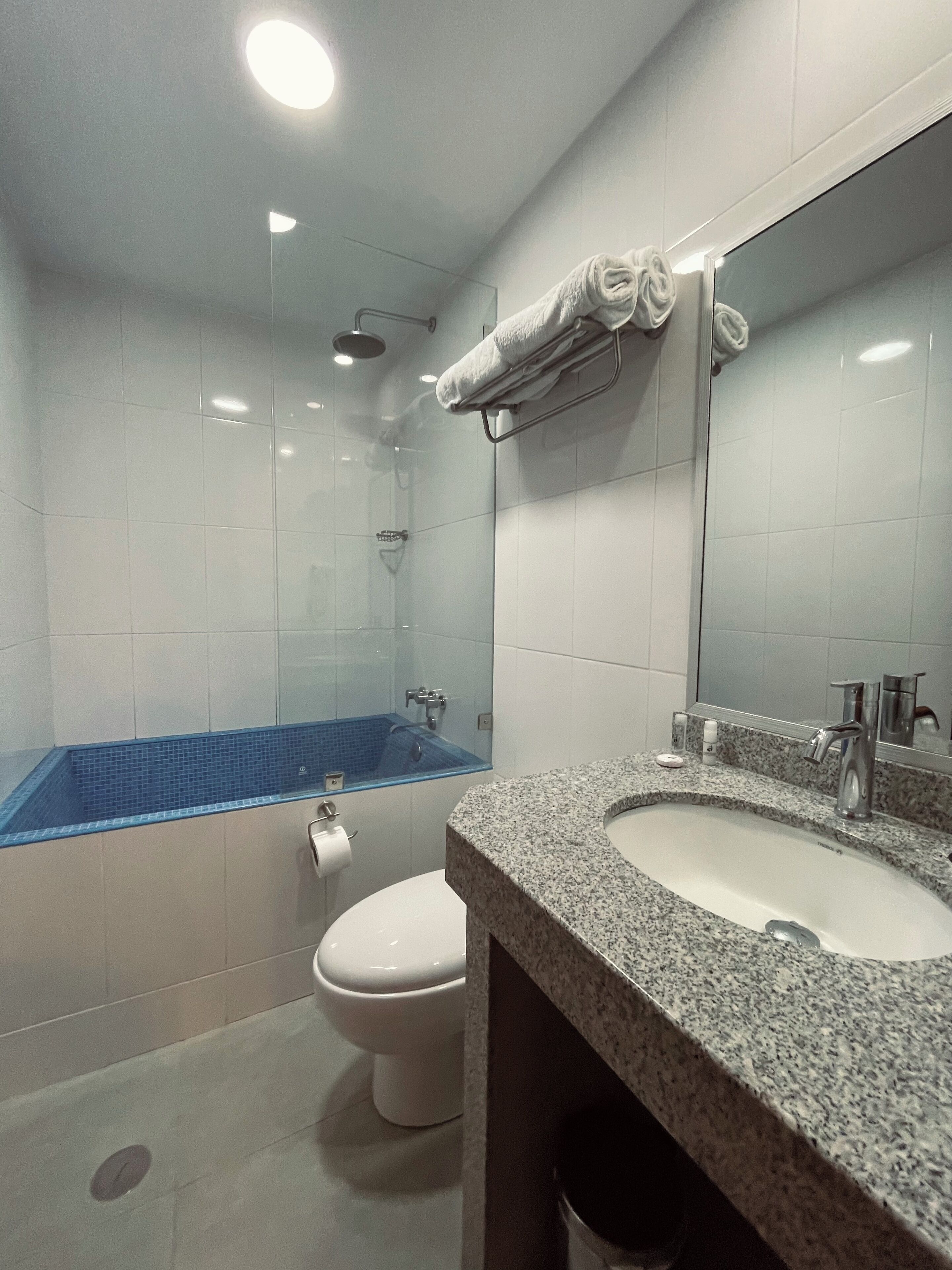 Un baño moderno con inodoro blanco, un lavamanos con encimera de granito, cabina de ducha de vidrio y un perchero para toallas.