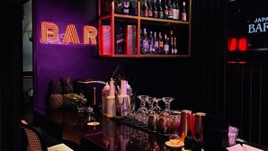 Un bar con un letrero de neón, estantes llenos de botellas y una variedad de utensilios y vasos de bar sobre la barra.