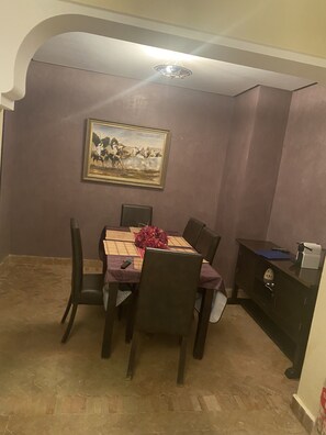 A dining area with a table set for four, a painting on the wall, and a desk with a lamp and a computer. - Un Joli Appartement Premium 23 (Marrakech)