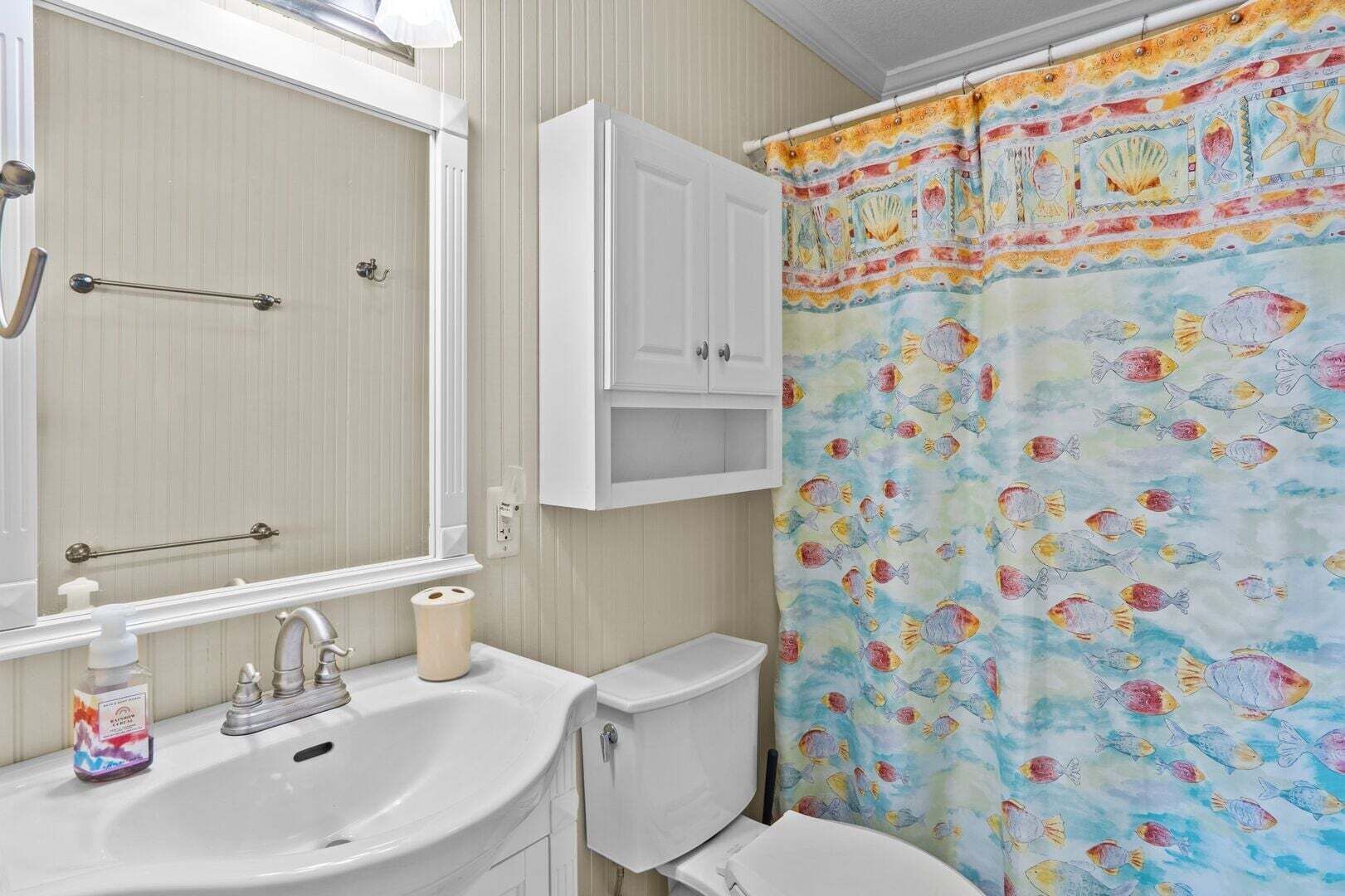 Une salle de bain équipée d’un lavabo, d’un miroir, de toilettes et d’un rideau de douche orné d’un motif de poissons.