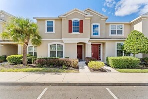 A two-story residential building with a red door, beige exterior, and a small front yard with a palm tree and shrubs. - 3 king suites + private heated pool, just steps to resort amenities. (Kissimmee)