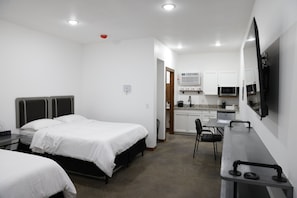 A compact hotel room with two single beds, a kitchenette, a dining area, and a flat-screen TV mounted on the wall.