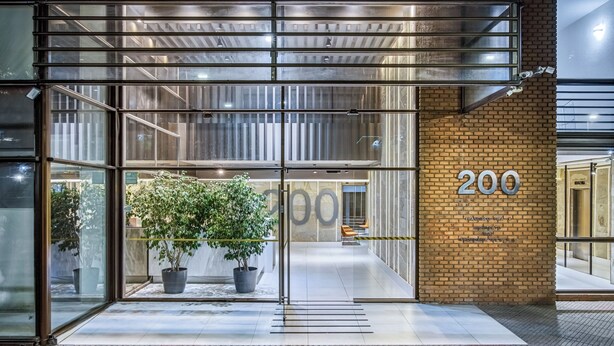 A modern building entrance with glass doors, a potted plant, and a brick wall with the number 200. - Studio apartment renewed @ best location in Santiago (Las Condes)