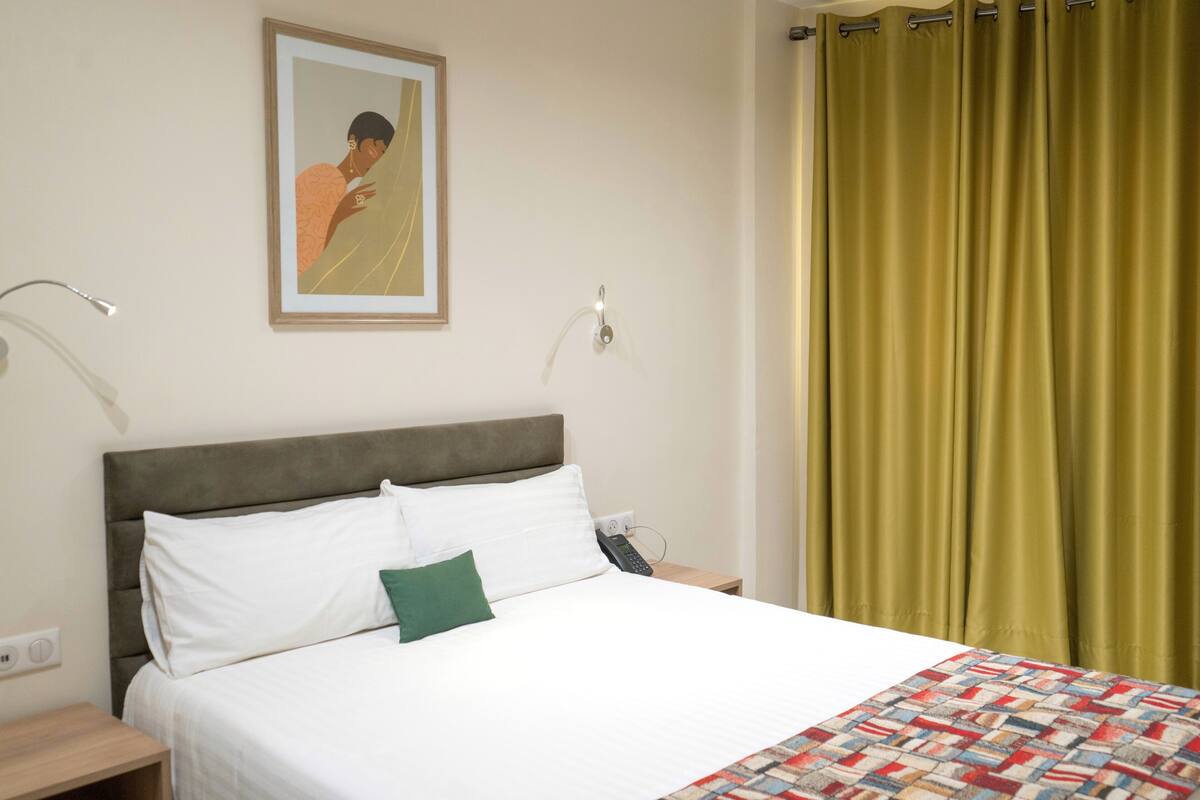 A hotel room with a bed, a nightstand, a painting, and a green curtain.