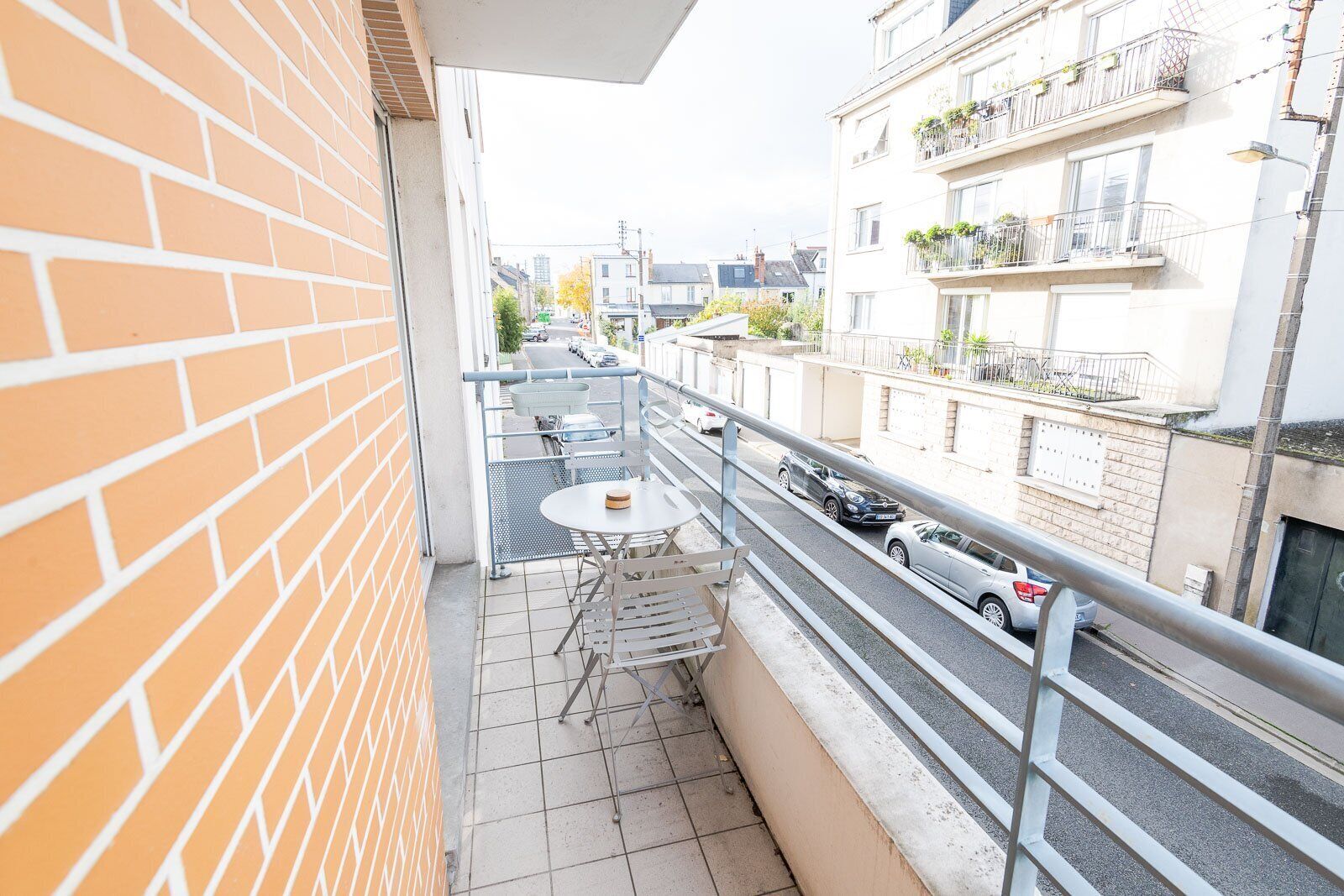 A balcony with a table and chairs, overlooking a street with parked cars and buildings.