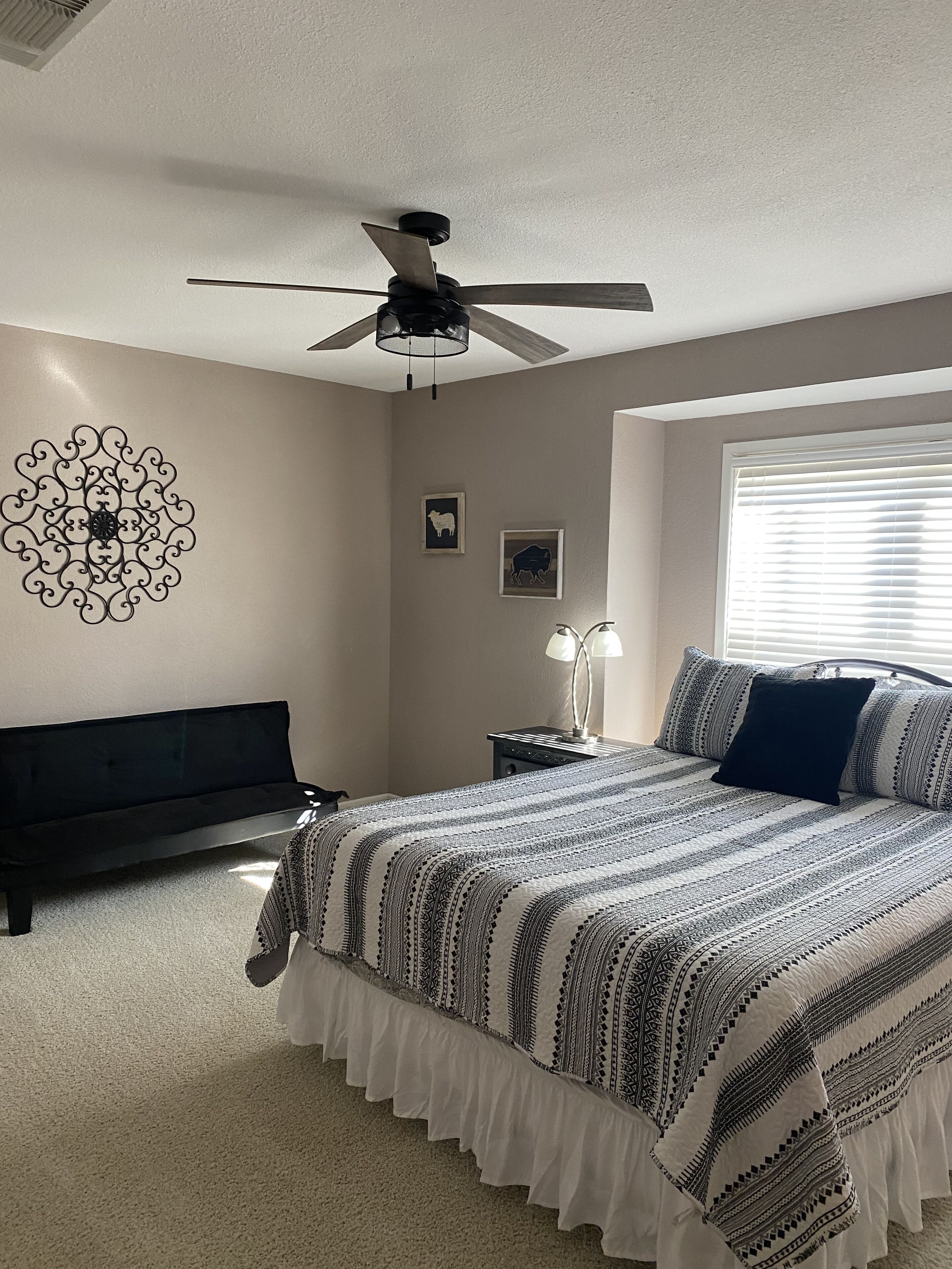 A bedroom with a bed, a ceiling fan, a wall decor, a window with blinds, and a bedside table with a lamp.