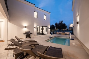 A modern outdoor pool area with lounge chairs, a barbecue, and a covered seating area.