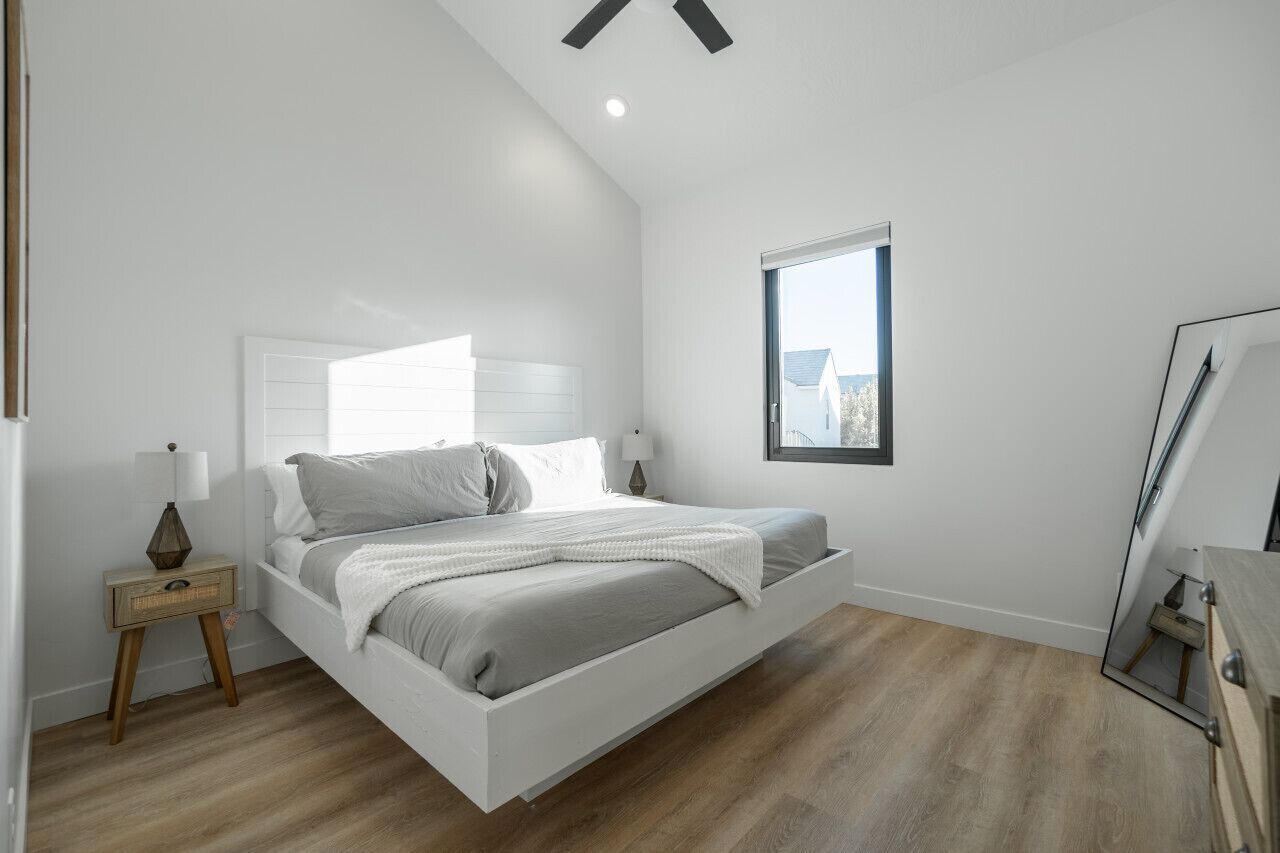 A modern bedroom with a white bed, a nightstand, a window, and a ceiling fan.
