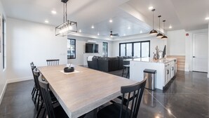 A modern kitchen with a central island, a dining area with a long table and chairs, and a living area with a sofa and TV.