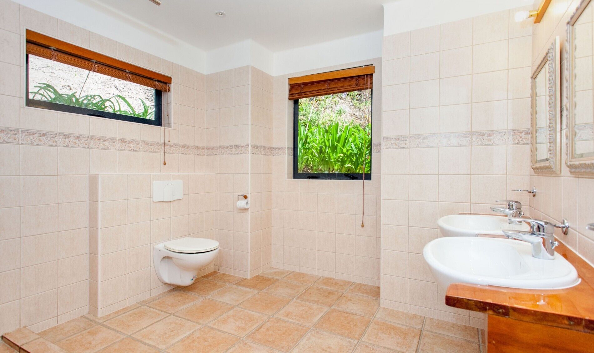 Un baño con inodoro, lavamanos y dos ventanas con vistas a zonas verdes.