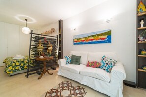 Una sala de estar con un sofá blanco, almohadas coloridas, un tapete estampado, un cuadro de barcos y un estante decorativo con figuritas.