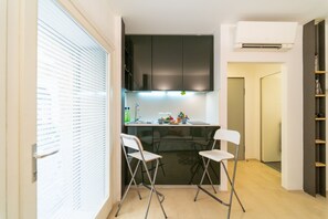 A compact kitchen with a dining area, a window with blinds, and a door leading to another room. - GuestHost - Ortigia Studios - Ground Floor Apartment (Siracusa)