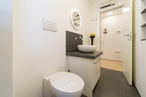 Un baño con inodoro blanco, lavamanos con pedestal negro, espejo y puerta decorada con un ancla.
