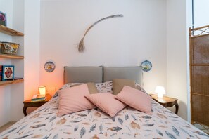 A bedroom with a bed, pillows, a headboard, a wall-mounted decorative item, a shelf with books, and a small table with a lamp.
