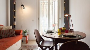 Un comedor con una mesa redonda, sillas y una lámpara. Se ve una ventana con cortinas y una puerta al fondo.