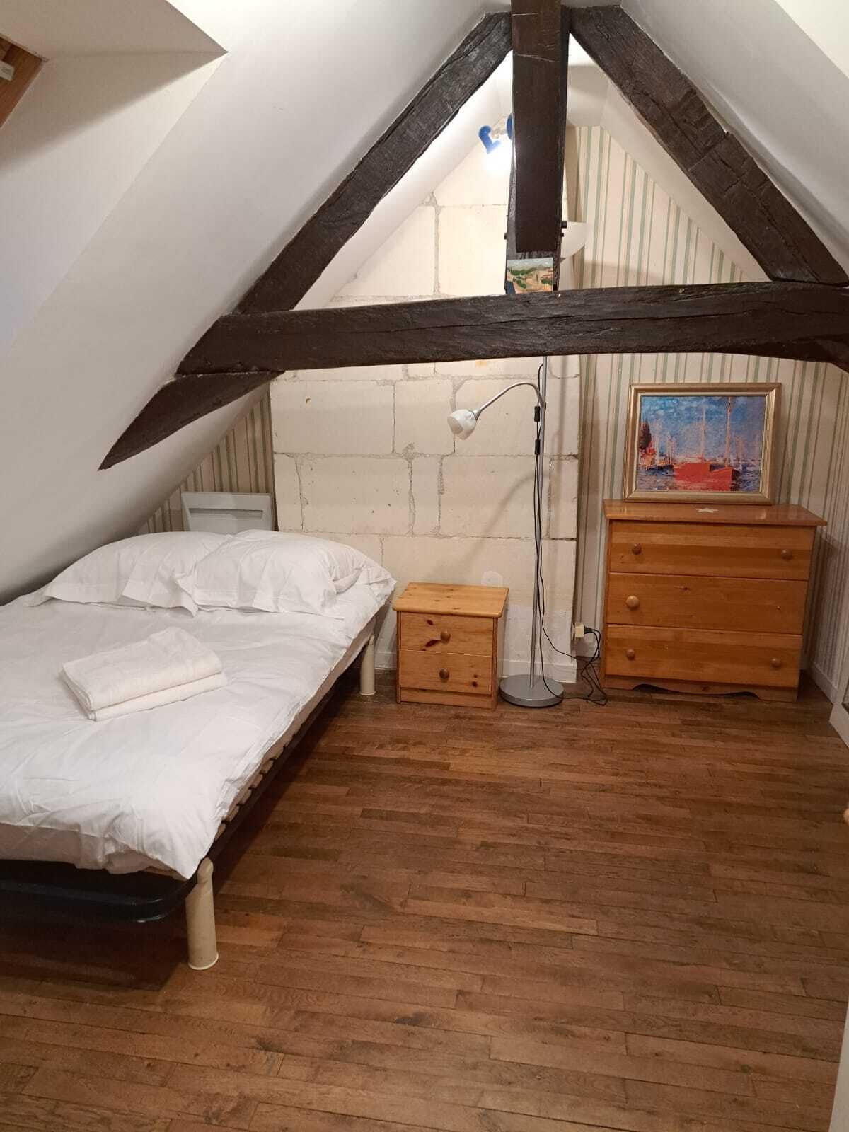 Een slaapkamer met een schuine plafond, een houten bed, een klein houten nachtkastje, een dressoir en een schilderij aan de muur.