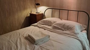 A single bed with white linens, a bedside table with a lamp, and a wooden wall panel.