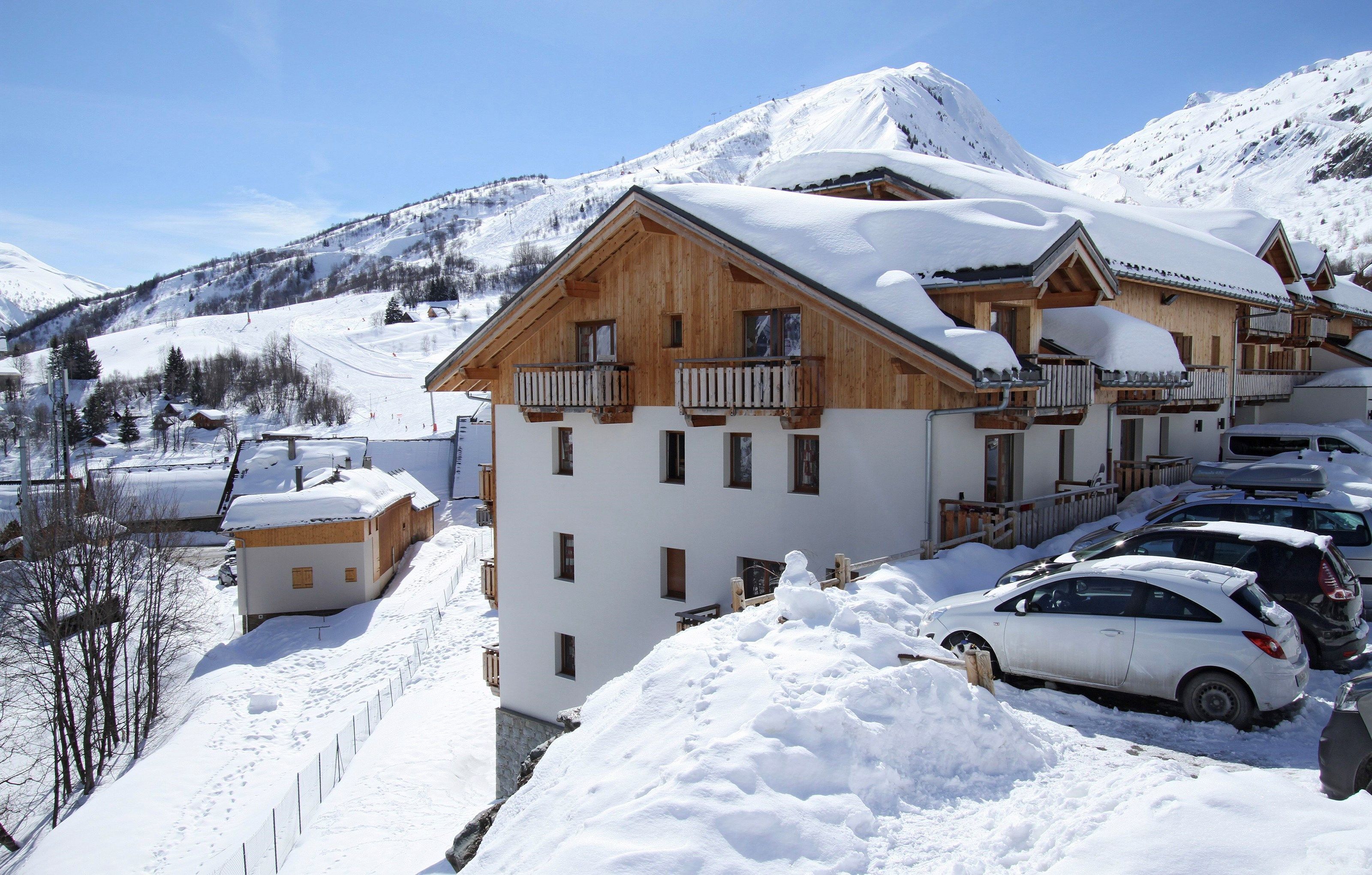 A wooden chalet with balconies, surrounded by snow-covered mountains and parked cars.