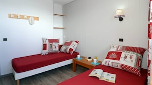 Una habitación con dos camas, cada una con ropa de cama roja y almohadas estampadas, una mesita de noche de madera con un libro y una taza, y una lámpara fijada a la pared.