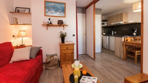 Un espace de vie compact comprenant un canapé rouge, une table basse avec une boisson et un livre, un coin cuisine avec des placards en bois, un four à micro-ondes et une table à manger.