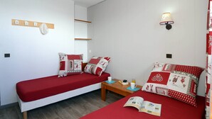 Une chambre avec deux lits, chacun agrémenté de draps rouges et d’oreillers à motifs, une table de chevet en bois avec un livre et une tasse, et une lampe fixée au mur.