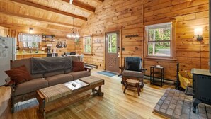 Ein Holzblockhaus-Interieur mit Holzboden, einer Couch, einem Sessel, einem kleinen Tisch, einem Herd und einer Küchenzeile mit Kühlschrank und Schränken.