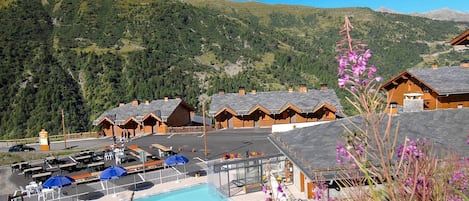 A mountain resort with a swimming pool, wooden chalets, and a view of the surrounding hills.