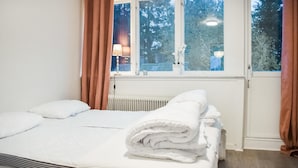 Un lit bien fait, avec du linge de lit blanc, un radiateur sous la fenêtre et une vue sur les arbres à l’extérieur.