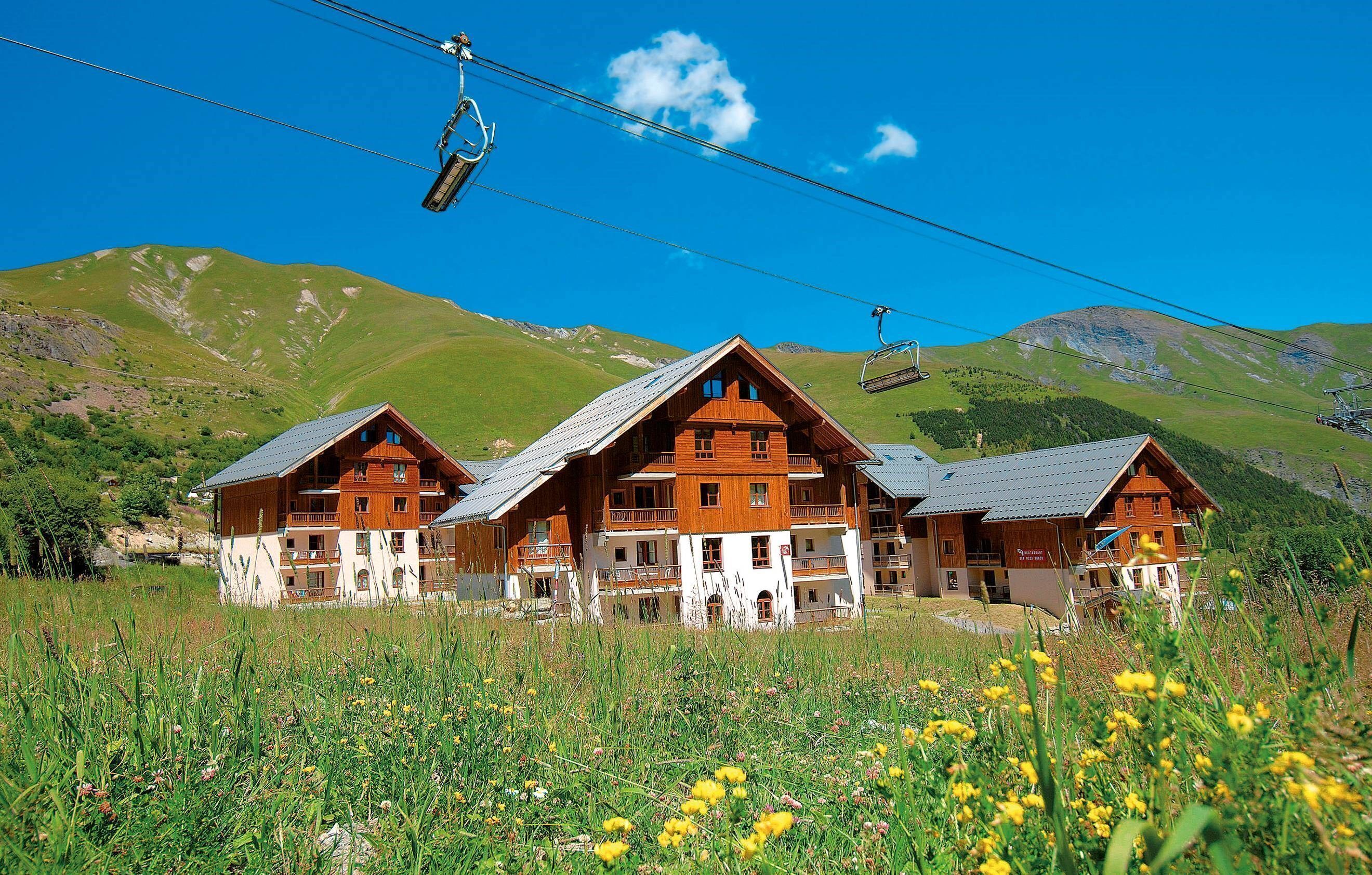 A wooden chalet-style building with multiple balconies, surrounded by a grassy field with yellow flowers, and a mountain backdrop with a ski lift in the distance.