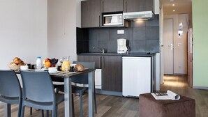 Eine kompakte Küchenzeile mit Mikrowelle, Kaffeemaschine und kleinem Kühlschrank. Ein Esstisch mit Stühlen und ein Frühstücksbuffet mit Croissants, Obst und Saft.