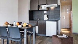 Cocina compacta con microondas, cafetera y un refrigerador pequeño. Cuenta con una mesa de comedor con sillas y preparación para desayunar.