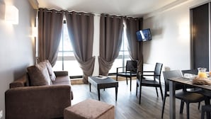 Une chambre d’hôtel moderne équipée d’un canapé, de deux fauteuils, d’une petite table avec des articles pour le petit-déjeuner et d’un téléviseur à écran plat fixé au mur.